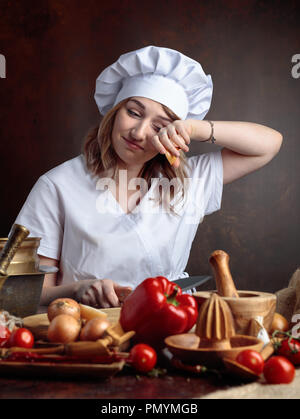 Young beautiful girl in a chef uniform cuts onions and cries. On the table different kitchen utensils and vegetables. Stock Photo