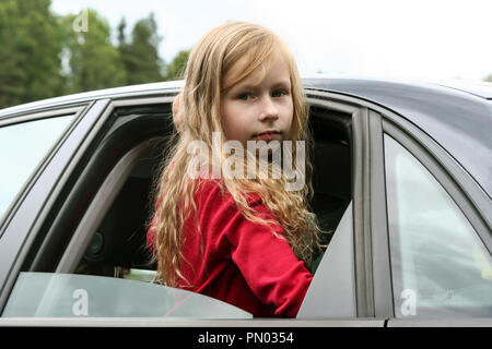 little girl with long blond hair and in a red jacket looks through the open window of the car into the camera, climbed out of the window to the waist, Stock Photo