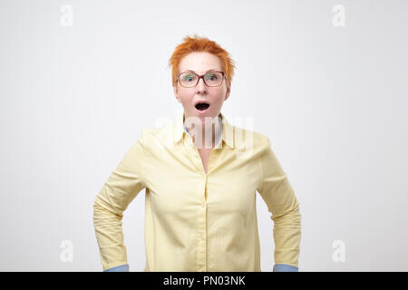 Oh no concept. Headshot of attractive european woman with glasses dressed in yellow shirt looking at camera, keeping mouth wide opened feeling shocked Stock Photo