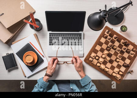 partial view of male freelancer with eyeglasses sitting at table working table with coffee, laptop and chessboard Stock Photo