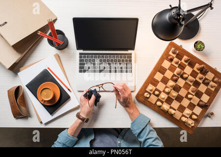 cropped image of male freelancer wiping eyeglasses at working table with laptop, chessboard and coffee Stock Photo