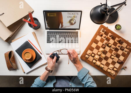 cropped image of male freelancer wiping eyeglasses at table with chessboard, coffee and laptop with tickets online on screen Stock Photo