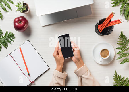 partial view of female freelancer using smartphone with blank screen at table with stationery, laptop and coffee surrounded by fern leaves Stock Photo