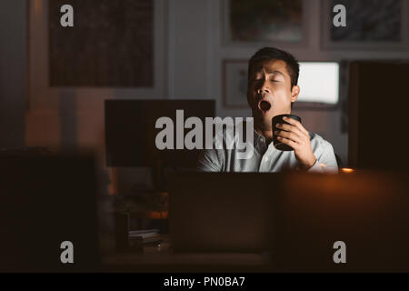 Young Asian businessman sitting at his workstation in an office late at night yawning while drinking a cup of coffee to stay awake Stock Photo