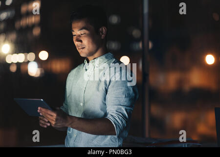 Young Asian businessman using a digital tablet while standing in an office late at night with city lights glowing in the background Stock Photo