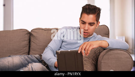 Young Hispanic man sitting on couch using tablet computer in living room Stock Photo