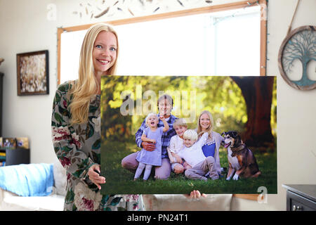 A happy young blonde woman is holding a large wall canvas portrait of her family with young children and a pet dog. Stock Photo