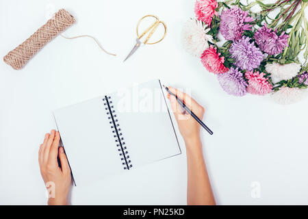 Female's hands writing in blank notepad among flat lay holiday border with copy space. Floral arrangement with bouquet of lush asters, twine and sciss Stock Photo