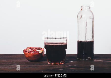 Freshly prepared pomegranate juice in glass next to half-empty bottle and cut fruit on dark brown wooden table on white wall background. Stock Photo