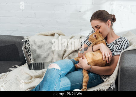 happy young woman holding red cat while using laptop at home Stock Photo