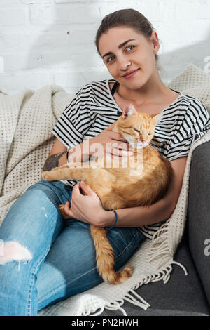 beautiful smiling young woman holding red cat and sitting on couch Stock Photo