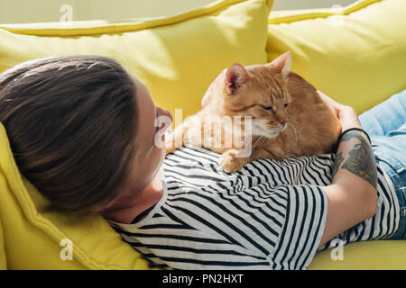 young woman holding cute red cat and lying on yellow couch Stock Photo