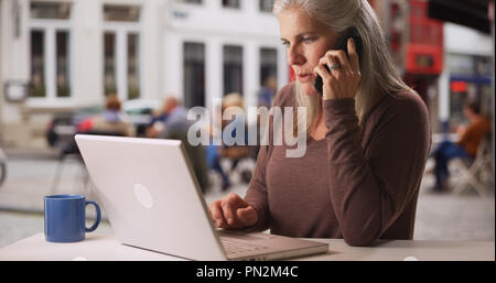 Busy older white woman works from laptop and makes phone call at cafe Stock Photo