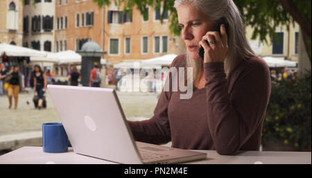 Caucasian female senior works on laptop at Italian cafe while talking on phone Stock Photo