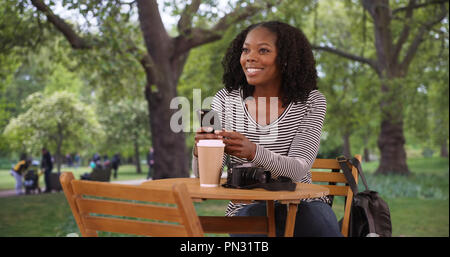 Portrait of pretty black woman sitting at table in city park holding phone Stock Photo