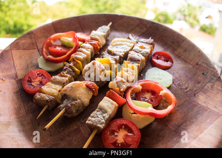 Skewered meat pieces prepared on grill with vegetables. Barbecued shish kebab or shashlik on sticks. Outdoor picnic meal on wooden round dish Stock Photo