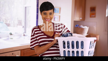 Portrait of cheerful black female holding laundry basket in kitchen laughing Stock Photo