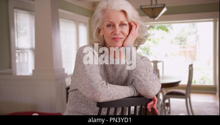 Lovely white female senior posing for camera sitting on chair inside nice house Stock Photo