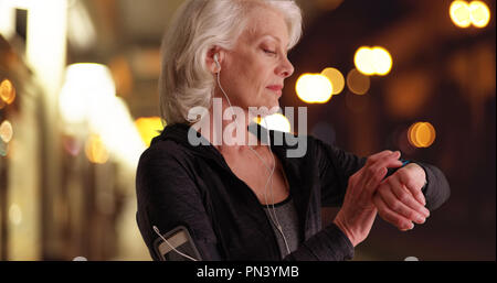 White athletic female elder checking watch while out jogging in city at night Stock Photo