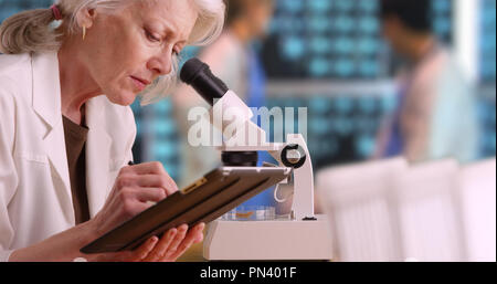Senior woman medical researcher taking notes on tablet computer in busy lab Stock Photo
