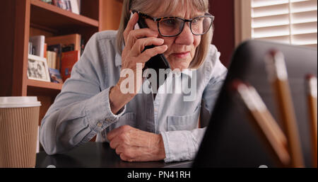 Close-up of old lady talking on phone and working on laptop in home office Stock Photo