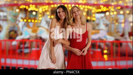 Two female best friends smiling and laughing near merry-go-round at street fair Stock Photo