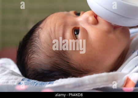 sweet little newborn asian baby drinking milk in plastic baby milk bottle relaxing on the bed and eyes contact to mother. Stock Photo