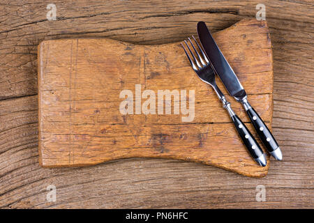 Knife and fork lie together with a very old cutting board on a rustic wood background Stock Photo