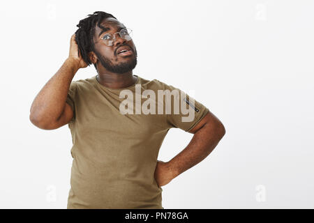 Waist-up shot of dreamy thoughtful dark-skinned man in glasses, scratching head and tilting up, looking upwards with hand on hip, thinking, being questioned and unsure while making plan Stock Photo
