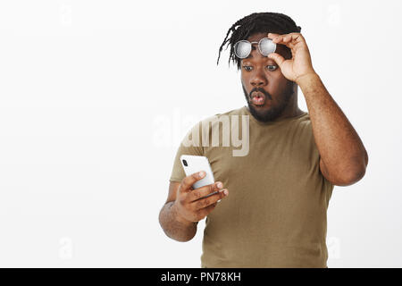 Guy whistling from surprise, being impressed with amount of money on account. Portrait of excited handsome plump african man, taking off glasses, holding rim on forehead and staring at smartphone Stock Photo