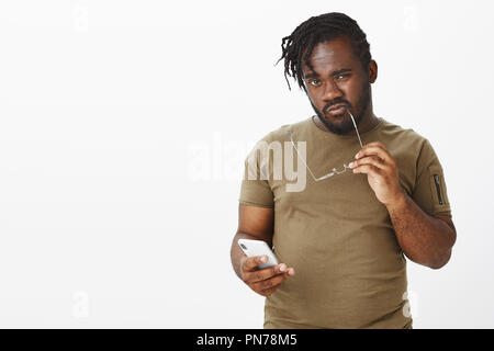 Waist-up shot of curious doubtful young african plump man in casual outfit, sucking rim of glasses and staring at camera with intrigued smart expression, holding smartphone, reconcireding plan Stock Photo