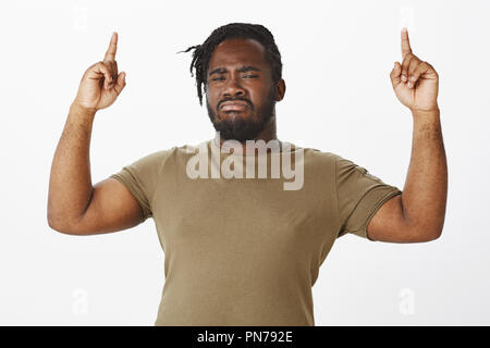 Waist-up shot of intense suspicious dark-skinned man in olive t-shirt, raising index fingers and pointing up with gloomy expression, being doubtful or hesitating while looking at new item in store Stock Photo