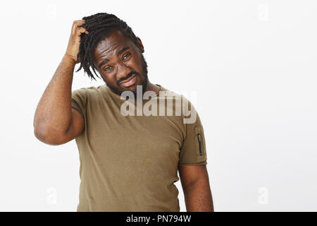 Waist-up shot of unsure and uncertain african-american guy scratching head and staring displeased at camera, being confused or stuck while making decision, having no idea what to do over grey wall Stock Photo