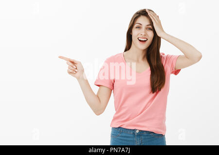 It totally slipped my mind. Portrait of confused happy good-looking woman in pink t-shirt pointing left with index finger while holding palm on head and smiling, being forgetful and asking question Stock Photo