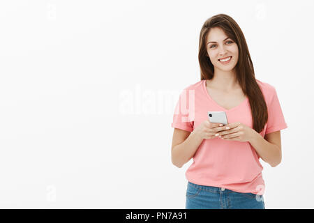 Studio shot of pleased charming woman in pink t-shirt, tilting head and smiling joyfully, holding smartphone over chest, tilting head while casually listening to interesting conversation Stock Photo