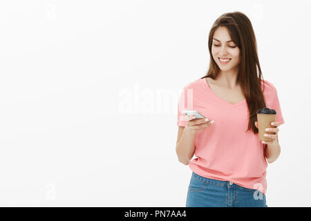 Girl talks with boyfriend via messages while he abroad. Portrait of good-looking happy nice woman in t-shirt holding cup of coffee and gazing at smartphone screen while reading interesting article Stock Photo