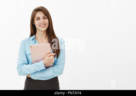 Positive attractive woman in office blouse, holding digital tablet with crossed hands on chest, smiling broadly, being friendly and polite, staring working at new place, meeting coworkers Stock Photo