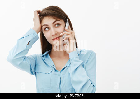 Girl not sure if she will be free. Confused good-looking and carefree female in blue blouse, scratching head and looking up with broad smile, talking on smartphone, making decision or thinking Stock Photo