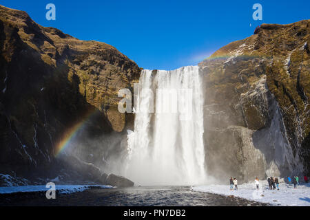 Tourists and rainbow at spectacular Skogar waterfall - Skogarfoss - in South Iceland with gushing glacial melting waters Stock Photo