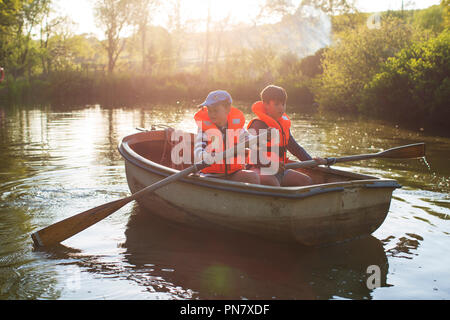 Two boys rowinig a boat on a small lake back lit by the evening sun. Stock Photo