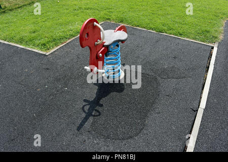 Red rocking bird piece of play equipment on a blue spring surrounded by a rubber crumb safety surface which has been repaired in several places and is Stock Photo