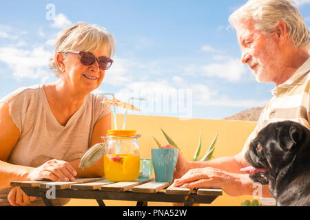 couple of aged gentlemen and lady drinkk together fruit juice on rooftop terrace wih two funny pug dog under the sunlight. amazing ocean view for grea Stock Photo