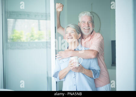 beautiful aged caucasian couple man and woman looking outside the window at home ina romantic moment of the day. foreverness life concept for caucasia Stock Photo