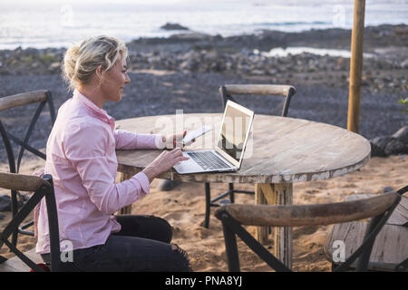 Young blonde business woman sitting outdoor near the ocean and the beach  and working with laptop and mobile phone hot sopt internet connection. alter Stock Photo