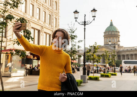 The young man waiting someone and does selfie, he dressed in a yellow sweater, a black raincoat or jacket, jeans, street and Kazanskiy cathedral Stock Photo