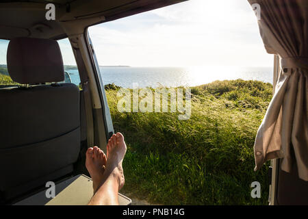 First-person view of a barefoot man relaxing inside a camper van and enjoying the view over the sea at sunset through the open sliding door. Stock Photo