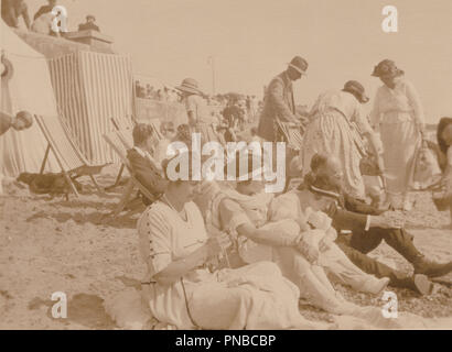* From The Outfits Possibly 1920's Photograph of People at The Seaside Sitting On The Sandy Beach. One of The Ladies is Knitting. Stock Photo