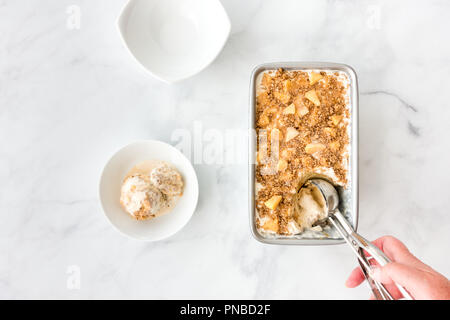 Hand scooping homemade ice cream with apples, caramel sauce and cinnamon crumbles out of a metal container. Stock Photo