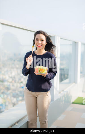 Portrait of attractive caucasian smiling woman eating salad on a sunny summer day sitting on green grass in park, focus on fork with salad Stock Photo