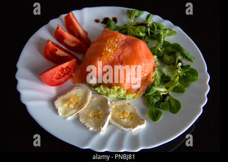 White plate with healthy meal on black background, in middle of plate is smoked salmon wrapped around avocado; topped with fish caviar, goat cheese Stock Photo